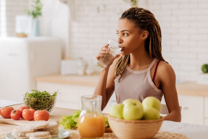 Femme dans sa cuisine en train de boire un verre d'eau et de manger des fruits et légumes frais, des gestes à adopter en cas de canicule