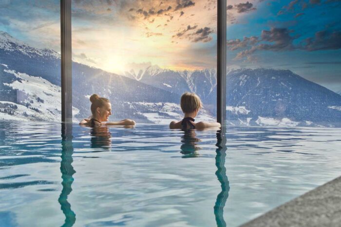 Deux femmes de dos dans une piscine intérieure avec une vue sur les Alpes