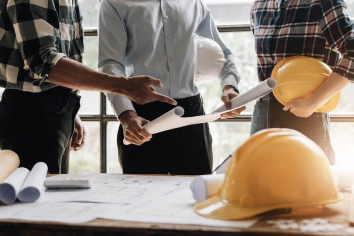 Trois personnes avec des plans de chantier dans les mains et des casques de chantier jaunes discutant